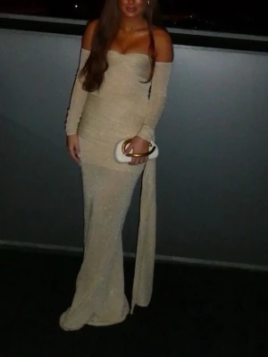 Lång glittrig offshoulder klänning - Säljer en elegant långklänning i ljus beige med glittrigt tyg. Klänningen har offshoulder-design, långa ärmar och figurnära passform. Perfekt för dig som vill sticka ut på bal eller fest. Inga synliga märken på plagget.