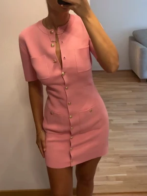 Oh polly  - Säljer en rosa kort klänning med guldknappar framtill och två stora fickor på bröstet. Klänningen har korta ärmar, rund halsringning och figurnära passform. Perfekt för dig som gillar en chic och trendig look.