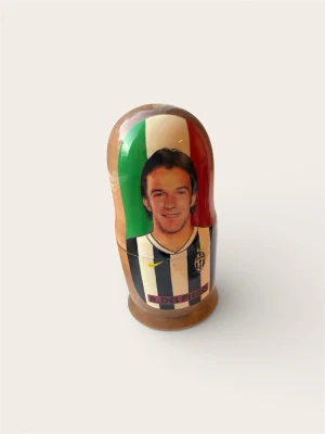 Alessandro Del Piero – Matryoshka / Rysk Docka (Juventus-souvenir) - Säljer en charmig och handmålad rysk docka (Matryoshka) föreställande Juventus-legendaren Alessandro Del Piero. Dockan visar Del Piero i den klassiska svartvita matchtröjan med italienska flaggan i bakgrunden.  En ultimat, lite udda inredningsdetalj för "Man Caven" eller en rolig present till den inbitne Juventus-supportern.  Skick: Fint skick! Lackerad yta utan synliga skavanker.  Mått: Standardstorlek för souvenir-dockor.