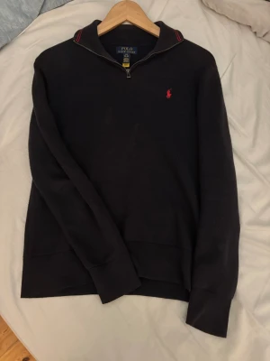 Mörkblå halvzip från Polo Ralph Lauren - Snygg mörkblå tröja från Polo Ralph Lauren med halv dragkedja och röd broderad logga på bröstet. Tröjan har ribbade muddar och krage med röda detaljer. Perfekt för en stilren och avslappnad look.