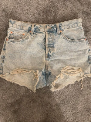 Ljusa denimshorts från One Teaspoon - Säljer ett par ljusblå denimshorts  med hög midja och slitna detaljer framtill. Shortsen har fransiga benslut, klassiska fem fickor och knappgylf. Perfekta för en avslappnad och trendig sommarlook.