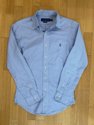 Blårutig skjorta från Ralph Lauren - Säljer en slim fit skjorta från Ralph Lauren i blåvitt rutigt mönster. Skjortan har klassisk krage, långa ärmar och knappar framtill. Ikoniska broderade loggan på bröstet. Tillverkad i 100% bomull för en skön känsla.