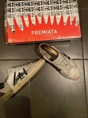 Vita och svarta sneakers från Premiata - Snygga vita sneakers från Premiata med svarta detaljer och grafiskt mönster på sidan. Skorna har klassisk snörning och perforerad tå för extra ventilation. Tillverkade i läder och kommer med originalbilden och kort och äktehetsbevis.