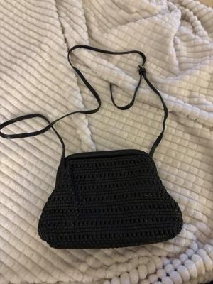 Svart virkad handväska med axelrem - Snygg svart handväska i virkat material med lång justerbar axelrem. Väskan har en unik formad botten och metallspänne upptill. Perfekt för dig som gillar en stilren och trendig accessoar med vintagekänsla.