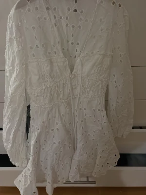 Vit broderad kortklänning från Zara - Söt vit kortklänning från Zara med broderade hålmönster och volangkant. Klänningen har v-ringning, långa ärmar och smock i midjan för en snygg passform. Perfekt för sommarens alla tillfällen!