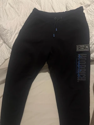 Svarta mjukisbyxor med tryck - Säljer ett par svarta mjukisbyxor med dragsko i midjan och blått tryck på ena benet. Byxorna har en modern passform och dragkedjeförsedda fickor på sidorna. Perfekta för chill dagar eller när du vill ha det bekvämt.