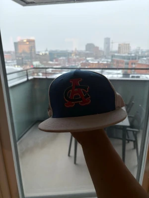 Blå och vit snapback keps med brodyr - Säljer en snygg snapback keps i blått och vitt med röd broderad logga på framsidan och ett färgglatt American Sports-märke på sidan. Platt skärm i beige och klassisk form. Perfekt för dig som gillar streetwear och sportig stil.