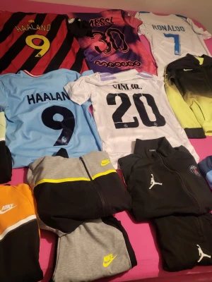 Fotbollströjor och Nike träningsset - Säljer flera fotbollströjor med spelarnamn som Haaland, Messi, Ronaldo och Vinicius Jr. Tröjorna har olika färger och tryck. Dessutom finns Nike och Jordan träningsset i svart, grått, gult och orange. Perfekt för dig som älskar fotboll och sportmode.