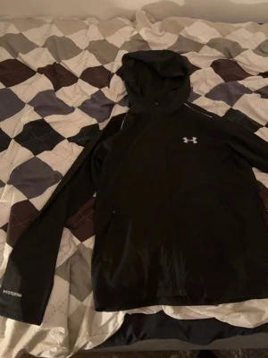 Svart vindjacka från Under Armour - Snygg svart vindjacka från Under Armour med huva och hel dragkedja framtill. Jackan har två sidofickor med dragkedja, reflekterande detaljer och logga på bröstet. Står storlek m i jacksn men passar mer som s