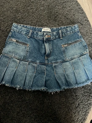Blå plisserad jeanskjol från Pull&Bear - Säljer en blå jeanskjol från Pull&Bear med plisserad nederdel och rå kant. Kjolen har två dragkedjefickor fram, bälteshällor och knappstängning. 