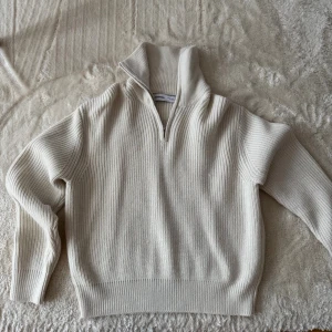 Beige stickad tröja från Bershka - Säljer en beige stickad tröja från Bershka med halv dragkedja och hög krage. Tröjan har ribbstickad struktur, långa ärmar och breda muddar vid ärmslut och nederkant. Perfekt för lager på lager och en clean look.