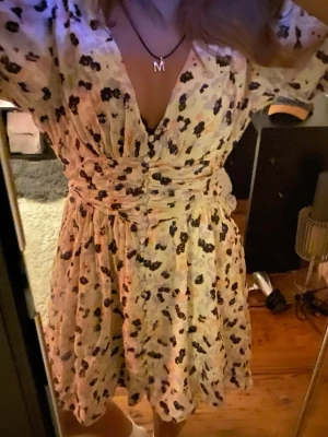 Blommig kortklänning med omlott - Söt kortklänning med blommigt mönster i svart och lila på ljus botten. Klänningen har omlottdesign, markerad midja och v-ringning. Trekvartsärm och lätt, luftig känsla. Perfekt för sommarens alla tillfällen.