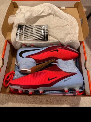 Fotbollsskor  - Säljer ett par Nike Phantom GT Elite AG-Pro fotbollsskor i rött och ljusblått med svarta detaljer. Skorna har strumpliknande skaft och är tillverkade i syntetmaterial för bästa grepp och kontroll på planen. Perfekta för konstgräs och avancerat spel.