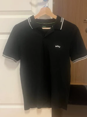 Svart pikétröja från BOSS - Klassisk svart pikétröja från BOSS med vit logga på bröstet. Tröjan har korta ärmar, krage med vita ränder och knäppning framtill. Perfekt för en stilren och sportig look.