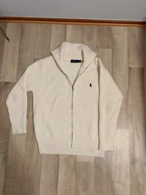 Vit ziptröja från Polo Ralph Lauren - Vit ribbstickad ziptröja från Polo Ralph Lauren med hög krage och dragkedja framtill. Tröjan har lång ärm och den klassiska broderade loggan på bröstet. Perfekt för lager-på-lager och chill stil.