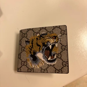 Gucci plånbok med tigertryck - Säljer en snygg Gucci-plånbok i canvas med klassiskt GG-mönster och ett stort broderat tigerhuvud på framsidan. Färgerna är beige, svart, gult och vitt. Plånboken har en fyrkantig form och känns riktigt lyxig och trendig.