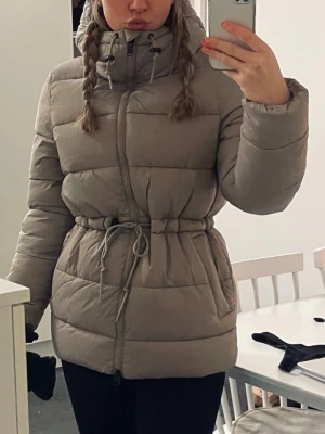 Beige pufferjacka med dragsko - Säljer en beige pufferjacka med hög krage, dragkedja och dragsko i midjan för snygg passform. Jackan har stora fickor och quiltad design som håller värmen. Perfekt för kalla dagar och riktigt skön att bära.