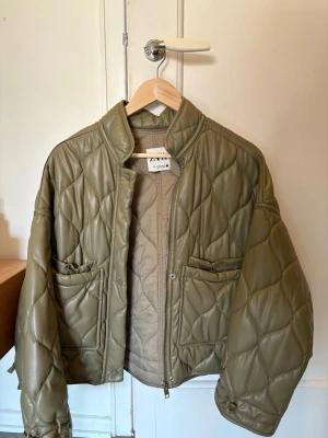 Olivgrön quiltad pufferjacka Zara - Säljer en olivgrön pufferjacka från Zara med quiltat mönster och två stora fickor framtill. Jackan har hög krage, dragkedja och tryckknappar. Materialet är i skinn. 