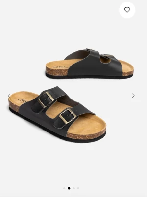 Svarta Linear sandaler - Klassiska svarta sandaler med två breda remmar och stora metallspännen. Perfekta för en avslappnad dag till stranden eller till vardags. Knappt använda. Ordinarie pris 349kr 