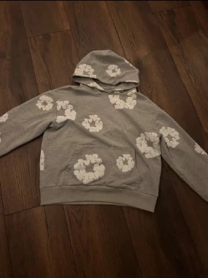 Grå hoodie med vita blommor Denim Tears - Snygg grå hoodie från Denim Tears med vita blommönster över hela tröjan. Klassisk huva och ribbade muddar. Perfekt för dig som gillar streetwear och vill sticka ut lite extra. Tillverkad i mjukt bomullsmaterial för skön känsla.