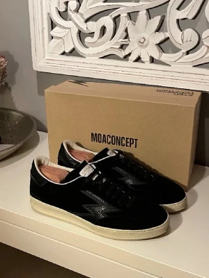 MOA Concept sneakers - Snygga svarta sneakers från MOA Concept med vit sula och vita detaljer. Skorna har en klassisk låg siluett, svarta skosnören och en ovandel i mocka och läder. Märkeslogga på plösen och sidan. Perfekta för dig som gillar stilrena och trendiga sneakers.