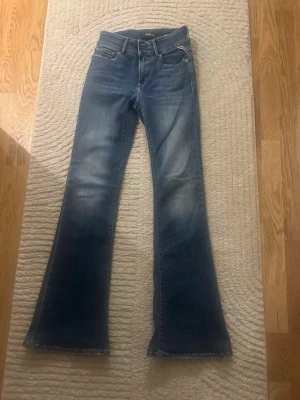 Blå utsvängda jeans från Replay - Säljer ett par blå utsvängda jeans från Replay, modell New Luz. Jeansen har klassisk femficksdesign, knappgylf och snygga slitningar framtill. Materialet är stretchigt denim som sitter bekvämt och följer kroppen. Perfekta för en trendig och avslappnad look.