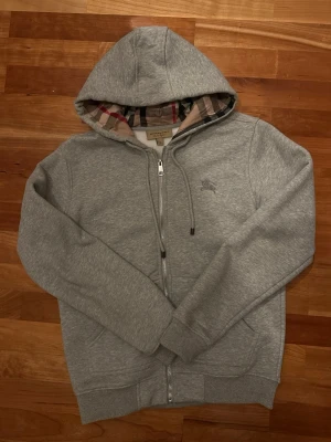 Grå zip hoodie från Burberry - Snygg grå hoodie från Burberry med dragkedja och klassiskt rutigt foder i huvan. Tillverkad i mjuk bomullsblandning, har två fickor fram och diskret Burberry-logga broderad på bröstet. Perfekt för en chill och stilren look.