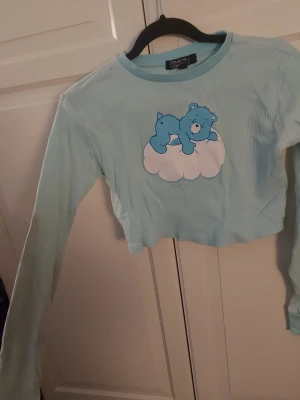 Ljusblå croppad topp med nalle - Supersöt ljusblå croppad långärmad topp från Dolls Kill med ribbad struktur. På bröstet finns ett tryck av en blå nalle som ligger på ett rosa moln. Perfekt för dig som gillar gulliga detaljer och vill sticka ut lite extra.