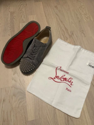 Grå Christian Louboutin sneakers - Grå sneakers från Christian Louboutin med klassisk röd sula och silverfärgade nitar på tån. En nit saknas längst ut på den vänstra skon som ni kan se på bilden. Utöver detta är skorna i bra skick. Dustbag inkluderas och box, äkthetscertifikat från vestiare finns även. Skorna är storlek 41 1/2 men sitter som 42.