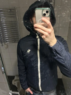 Mörkblå vindjacka från Abercrombie - Snygg mörkblå vindjacka med vit dragkedja och huva från Abercrombie & Fitch. Jackan har en ikonisk älg-logga på bröstet och är tillverkad i ett lätt, vindtätt material. Perfekt för blåsiga dagar och enkel att matcha med jeans.