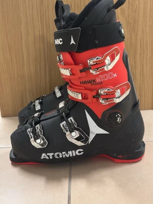 Atomic Hawx R100x Prime Pjäxor - Säljer ett par Atomic Hawx R100x Prime pjäxor i svart och rött med fyra metallspännen och robust plastskal. Sköna att åka i och ger bra stöd i backen. Snygg design med vita loggor och detaljer. Perfekta för dig som vill ta åkningen till nästa nivå.