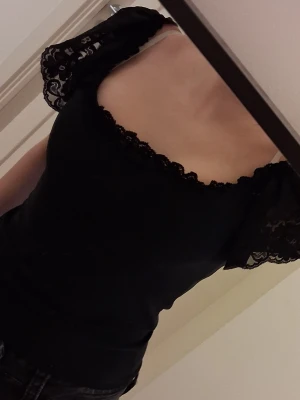 Spets topp - Snygg svart spets topp med spetskant runt urringningen och genombrutna spetsärmar. Modellen är figurnära och har korta ärmar med volang. Perfekt för dig som vill ha en feminin och trendig look.