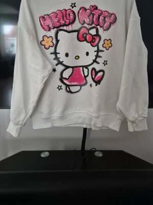 Vit Hello Kitty hoodie - Säljer en vit hoodie med stort Hello Kitty-tryck framtill. Motivet har rosa och gula detaljer, och texten 'Hello Kitty' i graffiti-stil. Hoodien har ribbade muddar vid ärmslut och nederkant. Den är även lite over sized i storleken.