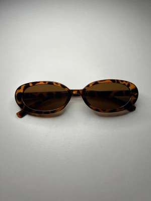 Ovala solglasögon i leopardmönster - Snygga ovala solglasögon med leopardmönstrad båge i bruna och svarta toner. Glasen är mörka och ger en cool retrovibe. Perfekta för dig som vill sticka ut med en trendig accessoar.