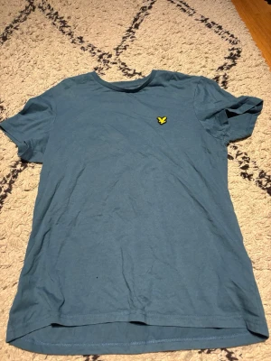 Blå t-shirt från Lyle & Scott - Säljer en blå t-shirt från Lyle & Scott med klassisk gul logga på bröstet. T-shirten har rund halsringning och korta ärmar. Perfekt basic-plagg med enkel och stilren design.