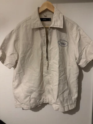 Vit Fortune Showrooms kofta - Kortärmad vit skjorta från Fortune Showrooms med krage och dragkedja framtill. Skjortan har tryckt logga på bröstet och stor logga på ryggen. Tillverkad i bomull och har en avslappnad passform som passar perfekt till en casual stil.