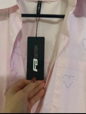 Ljusrosa skjorta med hjärtan FB Sister - Söt ljusrosa skjorta från FB Sister med glittriga hjärtan som detalj. Skjortan har klassisk krage och knäppning framtill. Perfekt för dig som vill ha en lekfull och trendig look.köpte för 299kr