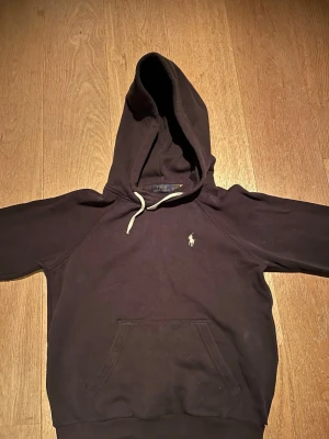 Svart hoodie från Polo Ralph Lauren - Snygg svart hoodie från Polo Ralph Lauren med klassisk vit logga broderad på bröstet. Hoodien har en stor huva med vita dragsnören och en praktisk magficka framtill. Tillverkad i mjukt bomullsmaterial som är skönt att bära.