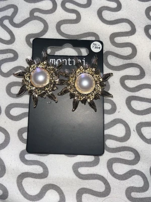 Guldiga örhängen med pärldetalj Montini - Statementörhängen från Montini i guldtonad metall med stora vita pärlor i mitten. Designen påminner om en sol med strålande detaljer runt pärlan. Perfekta för att lyfta vilken outfit som helst.