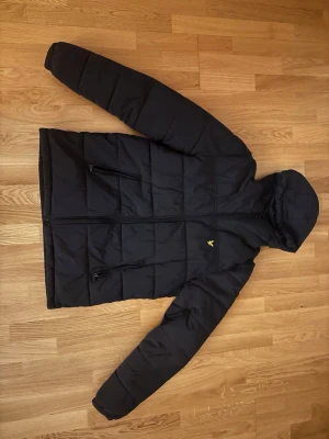 Svart pufferjacka från Lyle & Scott - Snygg svart pufferjacka från Lyle & Scott med huva och dragkedja framtill. Jackan har två snedställda fickor med dragkedja och den klassiska gula loggan på bröstet. Perfekt för kalla vinterdagar och riktigt skön att bära.