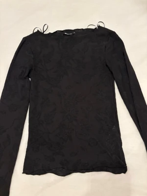 Svart blommig topp Gina Tricot - Snygg svart långärmad topp från Gina Tricot i transparent mesh med diskret blommigt mönster. Toppen har smala axelband med rosett-detaljer och sitter tight. Perfekt att styla med jeans eller kjol för en trendig look. 