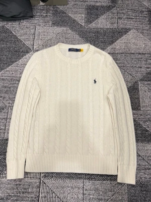 Vit stickad tröja från Polo Ralph Lauren - Kabelstickad vit tröja från Polo Ralph Lauren med rund halsringning och den klassiska broderade loggan i marinblått på bröstet. Tröjan har ribbade muddar vid ärmslut och nederkant. Perfekt för dig som gillar stilren och tidlös design. TTS