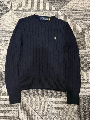 Mörkblå stickad tröja från Polo Ralph Lauren - Mörkblå kabelstickad tröja från Polo Ralph Lauren med rund halsringning och vit broderad logga på bröstet. Tröjan har långa ärmar och breda ribbade muddar vid ärmslut och nederkant. Perfekt för en stilren och klassisk look. NTTS