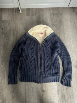 Mörkblå kabelstickad Levi's kofta - Mysig mörkblå kabelstickad kofta från Levi's med dragkedja framtill och vit teddyfoder på insidan. Stora ribbade muddar och broderad Levi's-logga i vitt på ryggen. Perfekt för kalla dagar när du vill vara både varm och snygg.