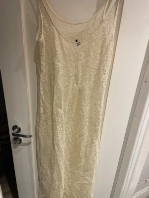 Lång beige stickad klänning - Säljer en lång, ärmlös stickad klänning i beige med rund halsringning. Klänningen har en enkel och stilren design som passar perfekt till sommaren. Materialet är stickat och ger en luftig känsla.