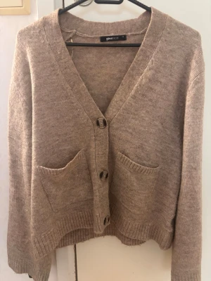 Beige kofta från Gina Tricot - Mysig beige kofta från Gina Tricot med v-ringning och tre stora knappar framtill. Koftan har två framfickor och ribbade muddar vid ärmslut och nederkant. Perfekt för lager på lager och enkel att matcha med olika outfits.