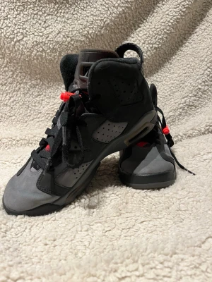 Svarta och gråa Air Jordan 6 sneakers - Grymma Air Jordan 6 psg sneakers i svart och grått med röda detaljer. Skorna har högt skaft, klassisk snörning och perforerade paneler. Ovandelen är i mocka och textil, sulan är svart med synlig luftdämpning. Perfekta för dig som gillar streetwear och basketstil.