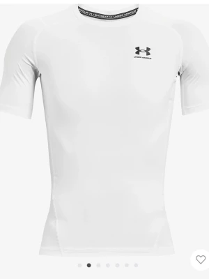 Vit kompressionströja från Under Armour - Säljer en vit, tight kompressionströja från Under Armour med korta ärmar och rund hals. Tröjan har svart logga på bröstet och textdetalj i halsen. Perfekt för träning och sportaktiviteter, tillverkad i ett stretchigt funktionsmaterial.