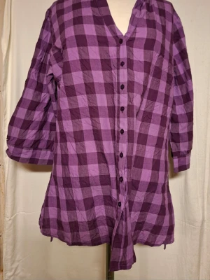 Lila rutig blus med knappar - Långärmad blus i två nyanser av lila med rutigt mönster och knappar framtill. Blusen har en avslappnad passform och v-ringad hals. Perfekt för dig som gillar färg och mönster. Stor medium