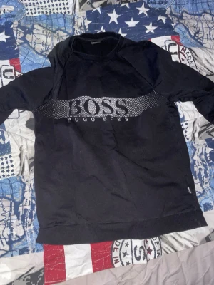 Svart huddie hugo boss - Snygg svart sweatshirt från Hugo Boss med stort logotryck över bröstet. Tröjan har rund halsringning, långa ärmar och ribbade muddar vid ärmslut och nederkant. Perfekt för en stilren och avslappnad look.